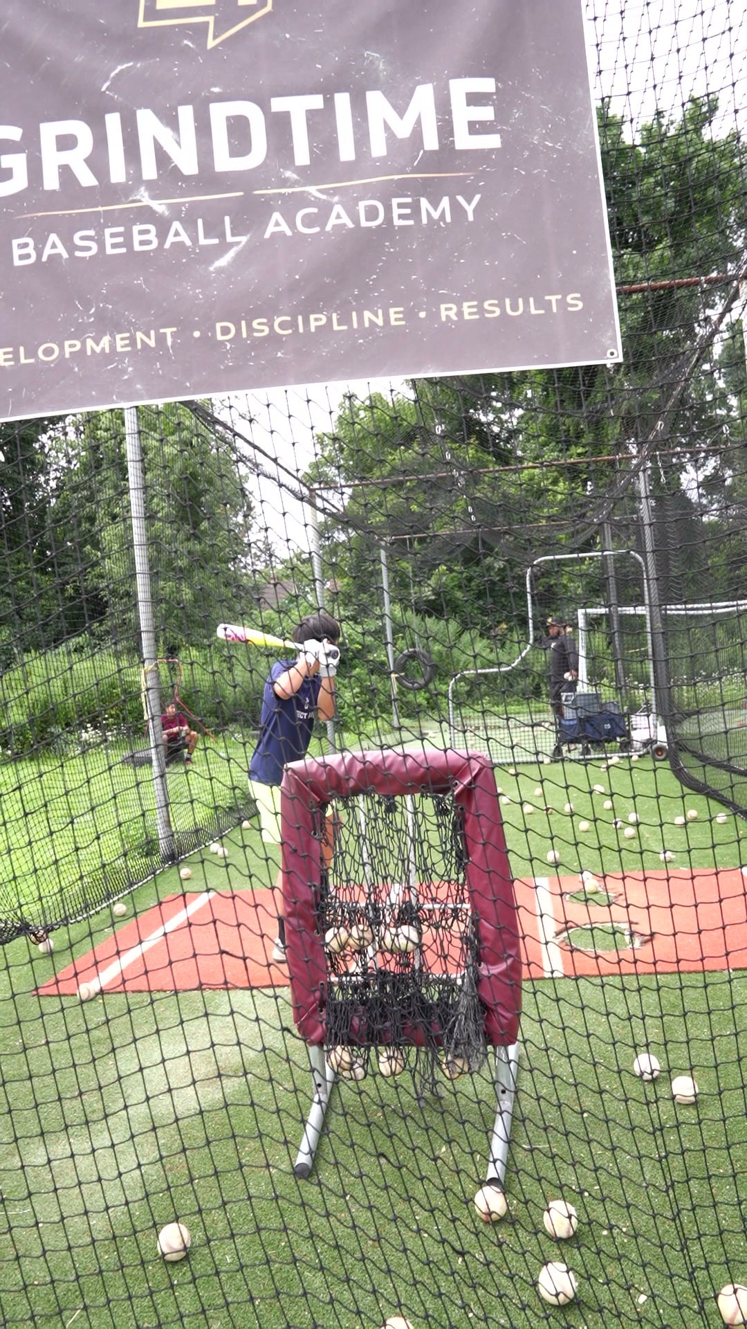 A young player swings in the GrindTime batting cage under the academy banner.