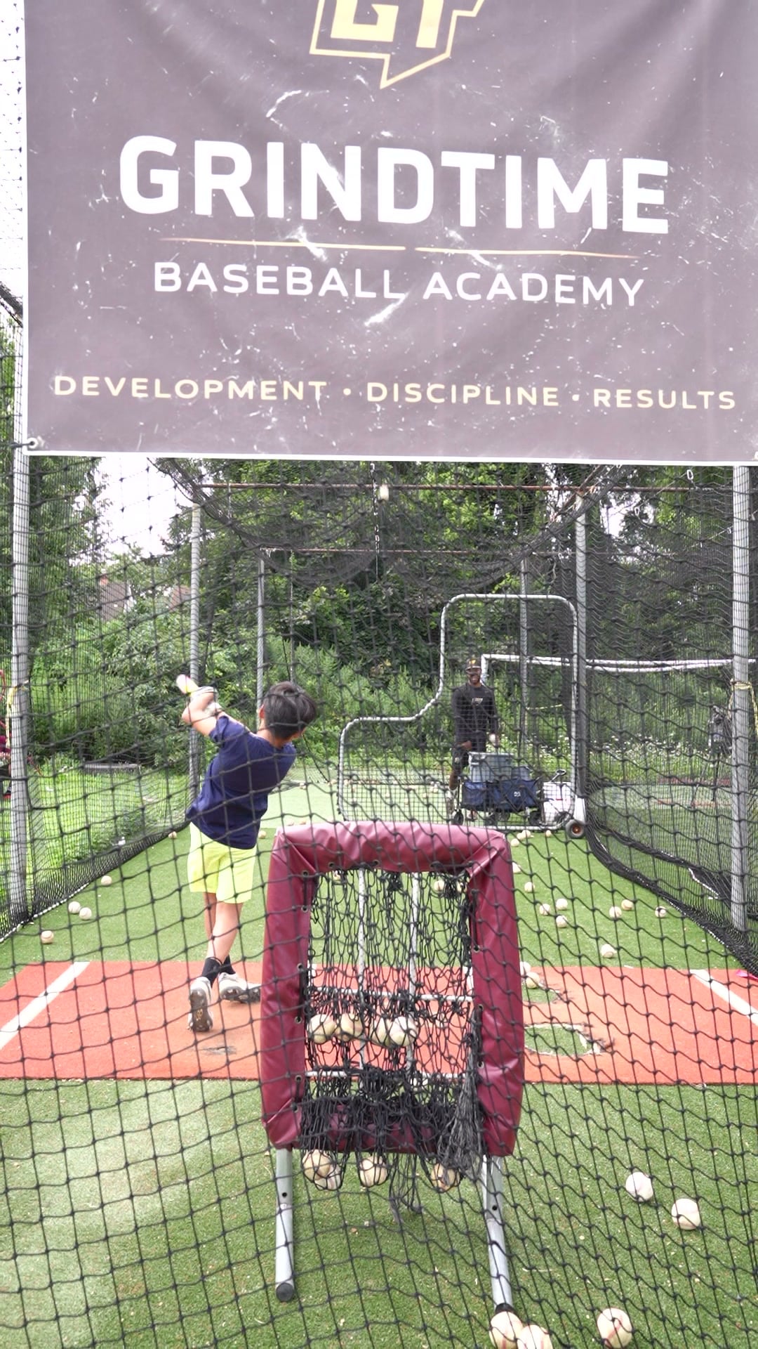 A player swings in the GrindTime batting cage under the academy banner.